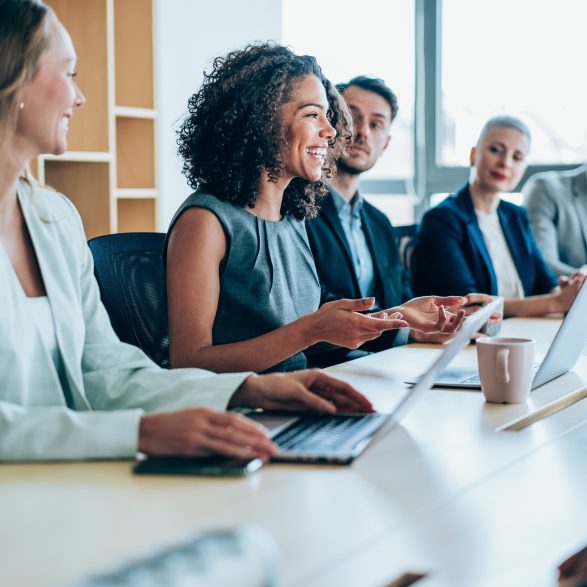 A business meeting in progress with people seated at a table with their laptops.