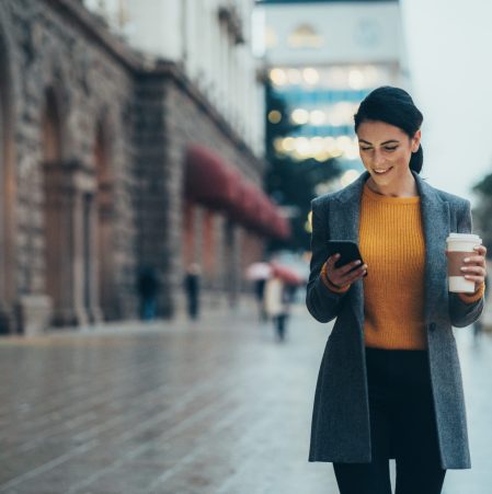 Modern young woman walking on the city street texting and holding cup of coffee