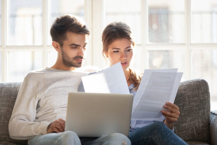 Millennial married couple sitting on couch with computer and documents, reading received formal letter from bank loan mortgage information, contract terms and conditions discuss papers working concept