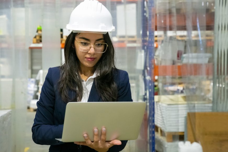 Focused female plant manager using laptop computer onsite. Middle aged woman in hardhat and uniform standing on plant floor. Production process monitoring concept