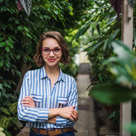 Young woman with arm crossed standing in botanical garden, looking at camera.