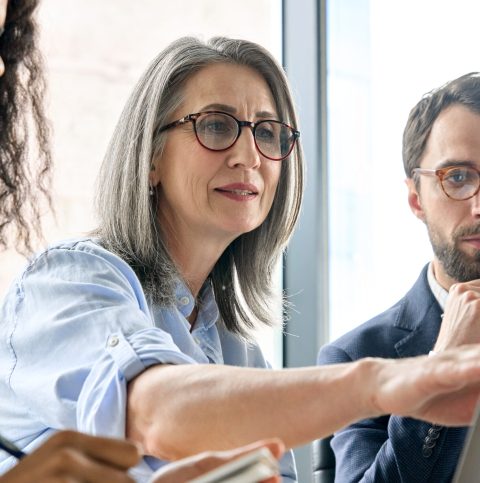 Mature older ceo businesswoman mentor in glasses negotiating growth business plan with diverse executive managers at boardroom meeting table using laptop. Multicultural team work together in office.