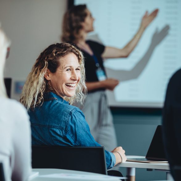 Rear view of a woman sitting in audience looking back at a colleague and smiling during a conference. Businesswoman smiling during a presentation.