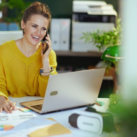 smiling modern 40 years old small business owner woman in yellow sweater with laptop speaking on a smartphone and working with documents in the modern green office.